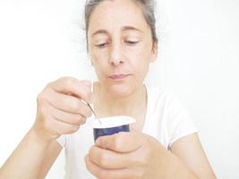 mujer de cuarenta y nueve años con una camiseta blanca sobre un fondo blanco comiendo un yogur foto