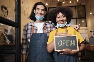 Two young startup barista partners stand at casual cafe door, write and show open sign before taking off face masks, happy and cheerful smiles with coffee shop service jobs after COVID-19 lifestyle. photo