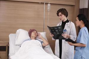 Caucasian female doctor in uniform diagnosis explains x-ray film with Asian radiologist and recovery male patient at inpatient room bed in a hospital ward, medical clinic, cancer examination consult. photo