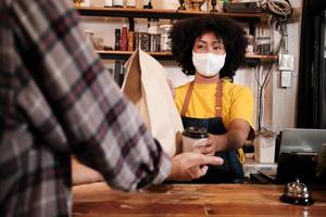 African American female barista with face mask works in the cafe with social distance, takeaway coffee for a customer, new normal service of small business coffee shop in COVID19 quarantine lifestyle. photo