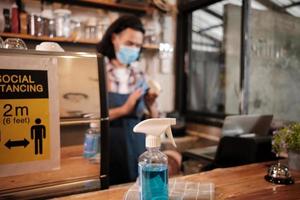 Alcohol sanitizer for healthy protection in a cafe, Asian male barista in face mask use laptop, waiting for coffee order in COVID19 pandemic quarantine, the economic impact for small business startup. photo