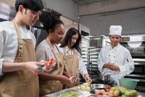 curso de cocina de hobby, chef masculino senior en uniforme de cocinero enseña a los jóvenes estudiantes de cocina a pelar y picar manzanas, ingredientes para pasteles, pasteles de frutas en la cocina de acero inoxidable del restaurante. foto