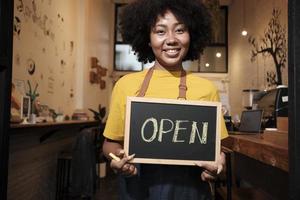 una barista nueva afroamericana se para en la puerta de un café informal, mira la cámara y muestra un cartel abierto, sonrisas felices y alegres con trabajos de servicio de cafetería y nuevos emprendedores de negocios. foto