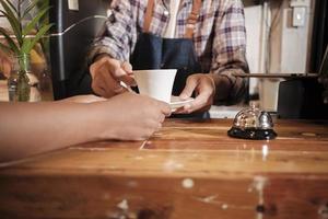 Front close-up of barista's hand works by serving white cup of hot coffee to customer order at wooden counter bar in casual cafe in morning, modern lifestyle works service job in coffee shop business. photo