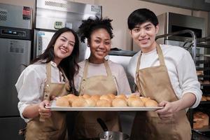 Portrait of three pastry startup partners looking at camera with a cheerful smile and proud with tray of bread in kitchen. A friend and founder of bakery foods and fresh daily bakery small business. photo