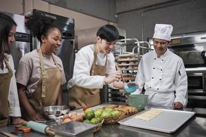 Cuisine course, senior male chef in uniform teaches young cooking class students, brushes pastry dough with eggs cream, prepares ingredients for bakery foods, fruit pies in stainless steel kitchen. photo