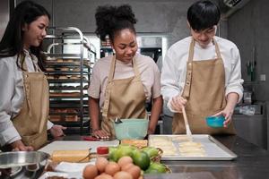 curso de cocina, un joven chef en delantal y un grupo de estudiantes de cocina, cepilla la masa de pastelería con crema de huevos, prepara ingredientes para alimentos de panadería, tartas de frutas en la cocina de acero inoxidable. foto