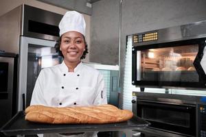 Portrait of African American female chef in white cooking uniform looking at camera with cheerful smile and proud with tray of baguette in kitchen, pastry foods professional, fresh bakery occupation. photo