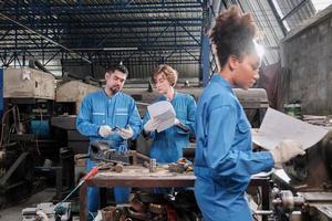 Two professional industry workers in safety uniforms and engineers' partners discussed mechanical drawing for lathe machines in manufacturing factory, with African American colleague in front of them. photo