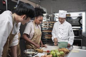 Cuisine course, senior male chef in cook uniform teaches young cooking class students to knead and roll pastry dough, prepare ingredients for bakery foods, fruit pies in stainless steel kitchen. photo