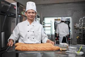 Senior Asian male chef in white cook uniform and hat showing tray of fresh tasty bread with a smile, looking at camera, happy with his baked food products, professional job at stainless steel kitchen. photo