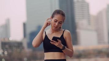 mujer joven en ropa deportiva con auriculares inalámbricos está descansando escuchando la música de su teléfono inteligente sosteniendo y cantando felizmente por la noche en el parque público. video