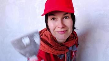 une femme drôle et positive dans une casquette et un uniforme de travail pose avec une spatule sur le fond d'un mur pour réparer et finir une pièce à la maison. réparer de vos propres mains, bricolage. attitude au travail video