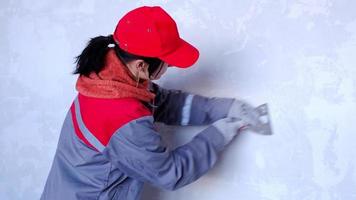 Preparation and cleaning of walls for repair and finishing with a spatula with plaster and putty before painting. A woman in a red cap and construction uniform is preparing the finishing of the walls video