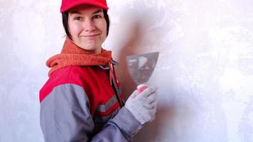 una donna divertente e positiva con un berretto e un'uniforme da lavoro posa con una spatola sullo sfondo di un muro per riparare e rifinire una stanza a casa. ripara con le tue mani, fai da te. attitudine al lavoro video