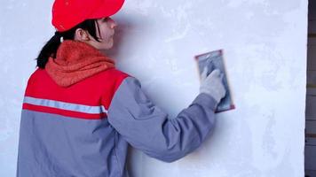 Sanding of walls with sandpaper after treatment with dry building mixes with plaster and putty before painting. A woman in a red cap and construction uniform is preparing the finishing of the walls video