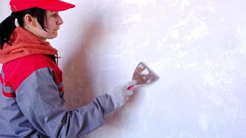 förberedelse och rengöring av väggar för reparation och efterbehandling med en spatel med puts och spackel före målning. en kvinna i röd mössa och bygguniform förbereder efterbehandlingen av väggarna video