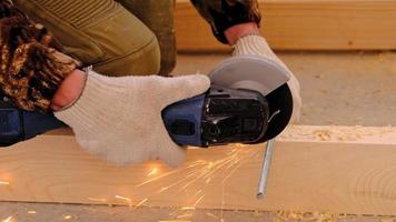 A builder's hands in work gloves cut a metal hairpin with an angle grinder electric tool. Preparation of a log for a wooden floor. Home repair with your own hands, a sawmill. Slow motion video