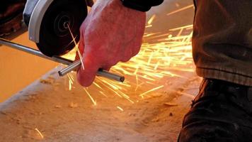 A builder's hands in work gloves cut a metal hairpin with an angle grinder electric tool. Preparation of a log for a wooden floor. Home repair with your own hands, a sawmill. Slow motion video