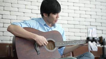 jovem asiático tocando violão e cantando uma música relaxe para gravar vídeo ao vivo, artista músico trabalha em casa, novo estilo de vida normal na comunicação on-line do ciberespaço video
