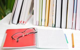 spiral copybook and eyeglasses on a table next to row of books and pencil. blank notebook. learning and study photo