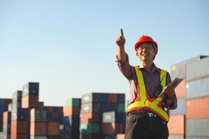 A senior elderly Asian worker engineer wearing safety vest and helmet standing and holding digital tablet at shipping cargo containers yard. photo