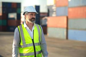 Caucasian male manager in white hard hat helmet and high-visibility vest walking in Container Terminal.