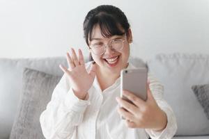 joven asiática que usa un teléfono inteligente para una videoconferencia en línea agitando la mano haciendo un gesto de saludo en el sofá de la sala de estar. foto