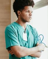Thoughtful young african american doctor standing by window looking away arms crossed. Portrait male doctor holds stethoscope seriously thinking, pondering. Professional medical occupation lifestyle.