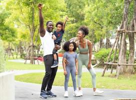 Happy family exercise in natural park. Black skin family, husband and wife have fun, laughing together with children at leisure in green garden. Playful kids have enjoyment outdoor with their parent photo