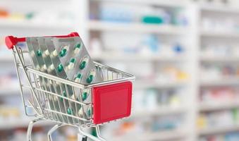 Medical pills capsule in shopping cart with pharmacy drugstore shelves blurred background photo