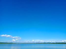 reflejo del cielo azul y la nube blanca en la superficie del lago con una línea de árboles y una pequeña montaña foto