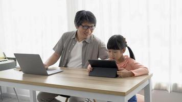 Happy Asian dad business man working from home and caring his daugther while watching funny cartoon on tablet in living room at home. Virtual homeoffice work and child care in family life. video