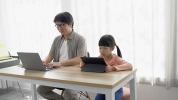 retrato de un padre asiático que trabaja desde casa con una computadora portátil y cuida a su niña viendo dibujos animados interesantes en una tableta digital mientras está sentado en el escritorio en casa. nuevo concepto normal. video