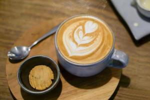 Top view of Late coffee and late art in blue ceramic coffee cup with a cookie in a black ceramic bowl on wooden plate and table in the coffee shop. photo