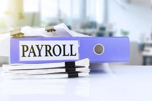 Payroll, the term is written on a folder of documents in trendy purple, lying on a stack of documents on an office table against the backdrop of an office with a soft blurred background.