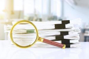 stack of paper documents with a magnifying glass on an office desk, against a blurred office background. Business and search concept. photo