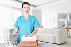 A beautiful young woman with an ironing board with an iron looks at the camera and smiles while ironing towels at home in the living room. Blurred background Home interior. Soft focus. photo