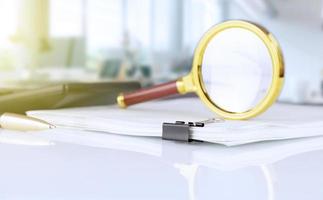 stack of paper documents with a magnifying glass on an office desk, against a blurred office background. Business and search concept. photo