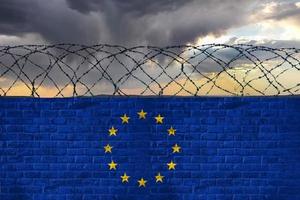 Brick wall with barbed wire in the colors of the flag of the European Union, EU, against a stormy sky. Crisis and hybrid war, border closures. photo