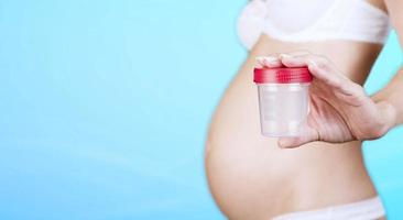 A pregnant Caucasian woman in underwear holding a plastic container for collecting urine, a jar with a red cap on a blue background. Close-up. Copy space. photo