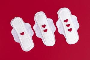 Three feminine sanitary pads with one, two and three red hearts on the feminine sanitary napkin. Feminine hygiene products during the menstrual cycle. Red background. Copy space. photo