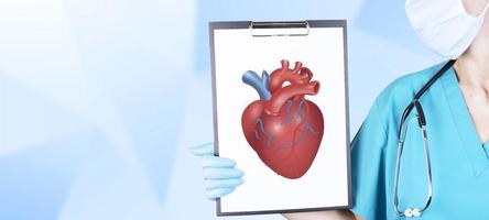 The hand of a cardiologist in medical clothes and a stethoscope holds a tablet with a realistic, voluminous, red heart on a light background. Cardiology concept. Copy space. photo