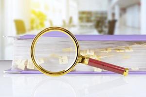 stack of paper documents in a purple folder with bookmarks and a magnifying glass on an office desk, against a blurred office background. Business and search concept. photo