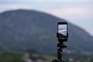 soporte de cámara de smartphone en un trípode, fotografiando una pintoresca puesta de sol sobre el mar tropical y las montañas. foto