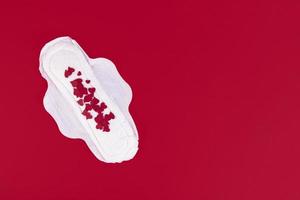 Many small red hearts, symbol of the menstrual cycle, on a feminine sanitary napkin. Feminine hygiene products during the menstrual cycle. Red background. Copy space. photo