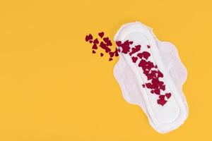 Many small red hearts, symbols of the menstrual cycle, on a feminine sanitary napkin. Feminine hygiene products during the menstrual cycle. Yellow background. Copy space. photo