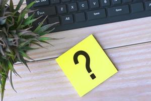 Question mark on adhesive note on wooden desk with keyboard and potted plant. photo