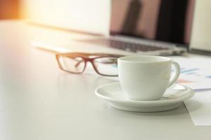 closeup coffee cup on table with laptop, glasses and papers in background, business concept. photo