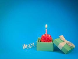 Sweet red cupcake with bow candle in gift box on blue background with copy space. Happy birthday party concept background. photo